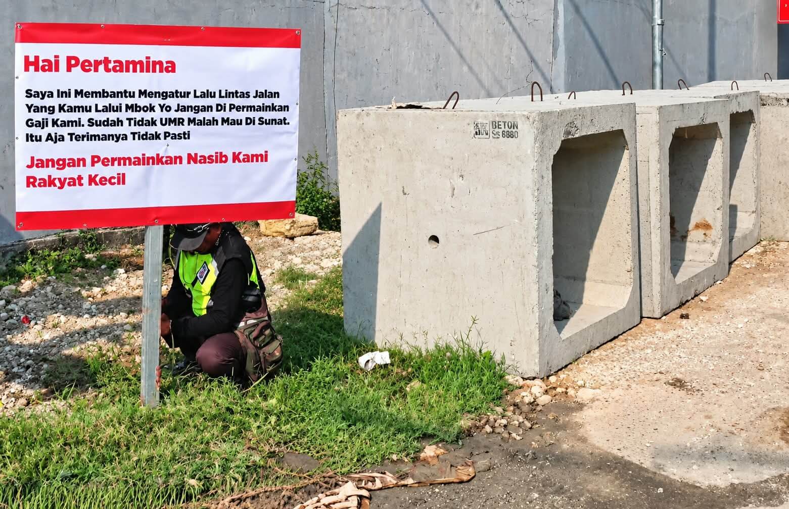 IMG-20261013-WA0072(1) Seorang flag man memasang spanduk berisi keluhan terkait gaji dan nasib pekerja pengatur lalu lintas di tepi jalan dekat pintu masuk area Pertamina.